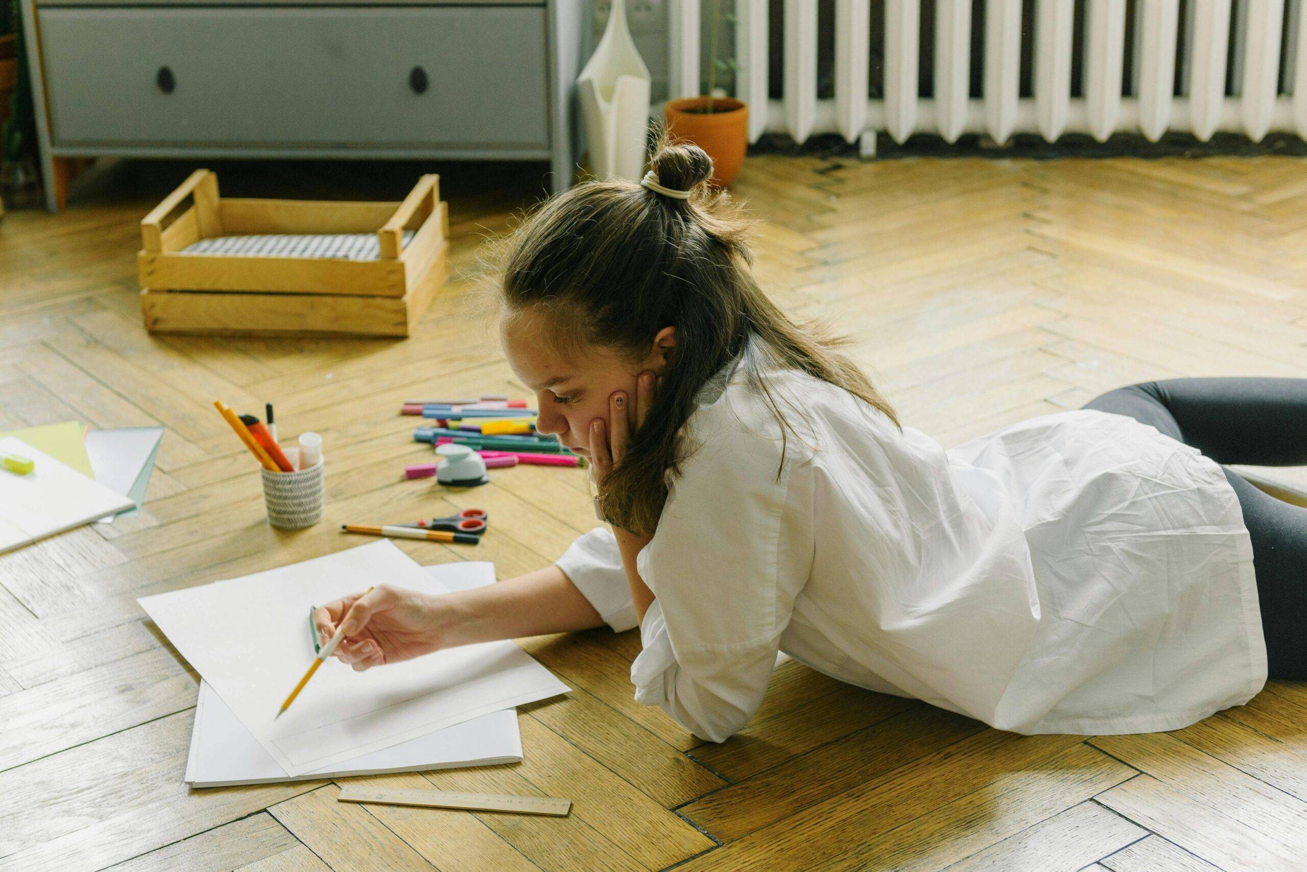 Teen girl draws on floor surrounded by art supplies, focusing on creative expression.